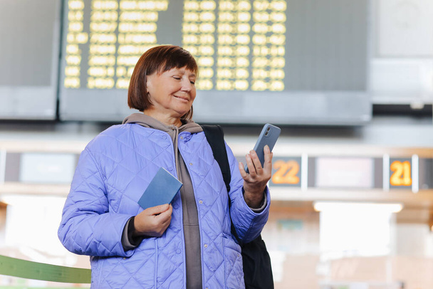 Keski-ikäinen nainen, jolla on reppu, pitää passia, käyttää matkapuhelinta lentokentän terminaalissa lähtöselvityksen jälkeen lentoyhtiön palvelupisteissä. Aktiivisia eläkeläisiä. Matkailu, loma, matkailukäsite - Valokuva, kuva