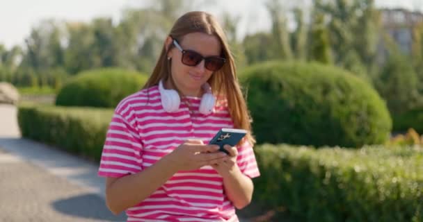 Una mujer atractiva y despreocupada que usa gafas de sol y auriculares alrededor de su cuello usa su teléfono celular durante un paseo pausado por un parque de la ciudad. Imágenes de alta calidad 4k - Metraje, vídeo