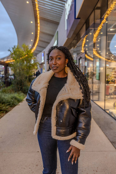 The photograph showcases a poised Black woman in an urban landscape during the evening. The setting sun casts a soft glow on the scene, illuminating her textured shearling coat and highlighting the - Photo, Image