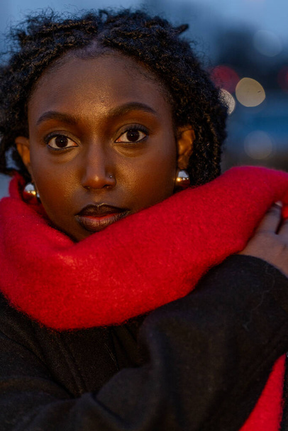 Het portret toont een verleidelijke zwarte vrouw gewikkeld in een levendige karmozijnrode sjaal, geplaatst tegen de schemertinten van de stad. Haar boeiende ogen zijn een brandpunt, het aantrekken van de kijker, terwijl de - Foto, afbeelding
