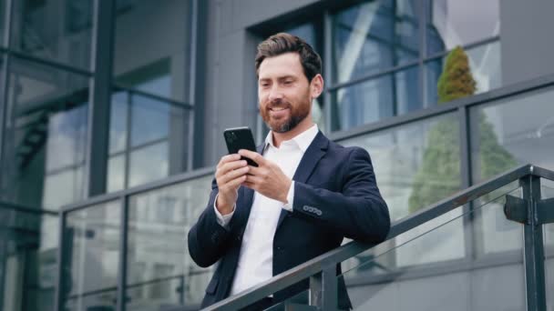 Udany uśmiech biały mężczyzna biznesmen lider przedsiębiorca za pomocą telefonu na zewnątrz w pobliżu biurowca przeglądanie biznesu social media sprawdzanie e-mail czat wysyłanie wiadomości na smartfonie - Materiał filmowy, wideo
