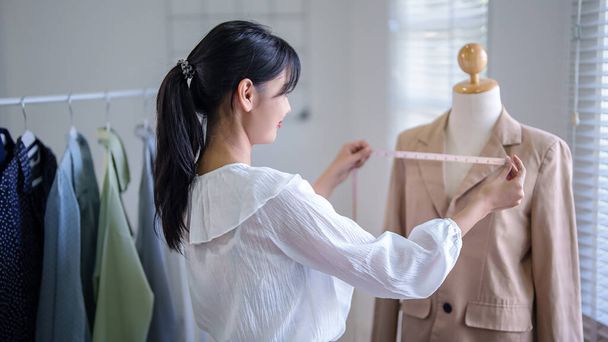 Νεαρές Ασιάτισσες έμποροι ελέγχουν τις λεπτομέρειες μεγέθους των νέων ρούχων μόδας σε μανεκέν με ταινία μέτρησης, ενώ κρατώντας σημειώσεις στο πρόχειρο και εργάζονται για online επιχείρηση αγορών στο γραφείο στο σπίτι. - Φωτογραφία, εικόνα