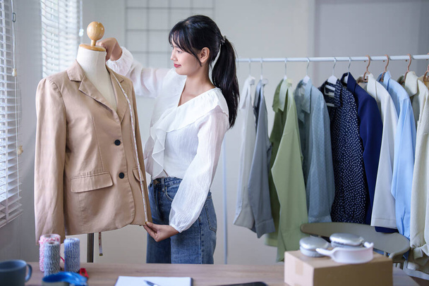 Młode azjatyckie kupieckie kobiety stosujące taśmę pomiarową do sprawdzania długości detali ubrań modowych w manekinach podczas robienia notatek w schowku i pracy nad zakupem online w biurze. - Zdjęcie, obraz