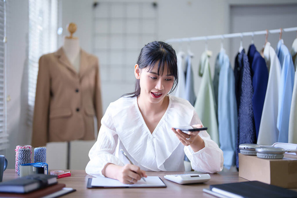 Nuoret aasialaiset kauppiaat naiset, jotka soittavat älypuhelimeen asiakkaan kanssa vaatetilauksen tiedoista ja kirjoittavat muistiinpanoja paperilla työskennellessään ja lähettäessään verkkokaupan toimitusta kotitoimistossa. - Valokuva, kuva