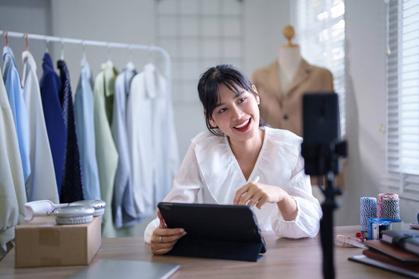 Jóvenes mujeres comerciantes asiáticas utilizan el teléfono inteligente para grabar video y vender productos de ropa en las redes sociales mientras trabajan para tomar notas en tabletas digitales sobre el negocio de compras en línea en la oficina en el hogar. - Foto, imagen