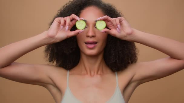 Schoonheid routine gelukkig jong meisje mooi aantrekkelijk Afro-Amerikaanse vrouw met krullend haar houden verse komkommer plakjes cirkels ogen dekking poseren beige studio achtergrond organische cosmetica huidverzorging - Video