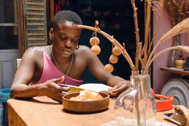 νεαρή Αϊτινή γυναίκα με κοντά μαλλιά στο σπίτι κάθεται στο σαλόνι διαβάζοντας άνετα, με κρουασάν σε καφέ κεραμικό πιάτο κύμα στο τραπέζι, έννοια του τρόπου ζωής, αντίγραφο χώρου. - Φωτογραφία, εικόνα
