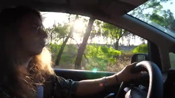 Profiili hippi mies aurinkolasit ajaa autoa auringonvalossa taustalla. Nuori hipsteri, jolla on pitkät hiukset ja joka ajaa autolla maaseudulle. Käsite matka ja matka. Sulje hitausliike. - Materiaali, video