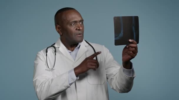 Pensive усміхнений афроамериканський чоловік чоловік радіолог лікар-хірург травматолог, який вивчає рентген в студії синій фон результати перевірки сканування рецензування частини тіла пацієнта медичний огляд - Кадри, відео