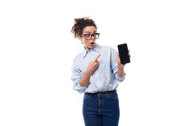 zaskoczony młody uśmiechnięty stylowy biały kobieta z kręconymi włosami demonstruje ekran smartfona z makietą. Koncepcja e-biznesu. - Zdjęcie, obraz