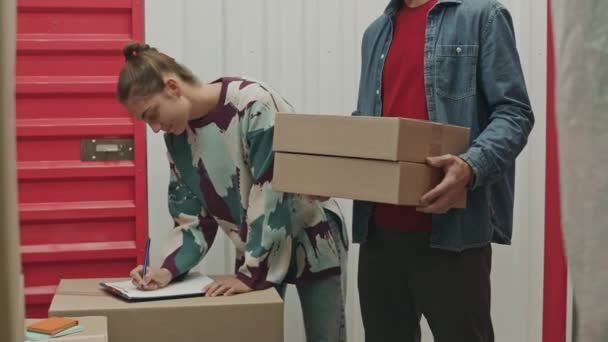 Blick aus dem Inneren der Self-Storage-Einheit einer Arbeiterin, die Notizen auf Klemmbrett macht und männlichen Kunden anweist, wo sie Kisten ablegen sollen - Filmmaterial, Video