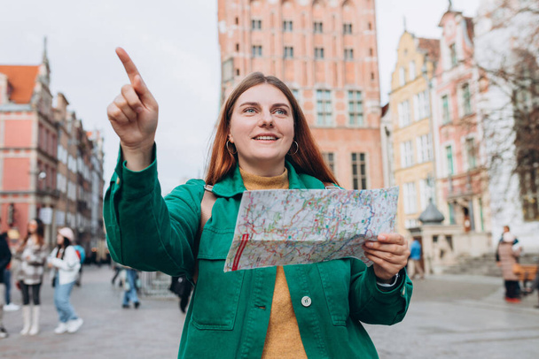 Portrait schöne Frau mit Papierkarte in der Straße. Glückliche Touristen reisen durch Europa. Rotschopfiges Mädchen mit Rucksack zeigt mit dem Finger auf die Straße. Europa im Herbst bereisen. - Foto, Bild