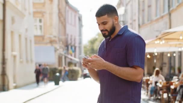 Turista viajante barbudo indiano homem usando smartphone digitando mensagens de texto à procura de uma maneira no mapa no aplicativo navegador móvel ao ar livre. Cara caminhando passa por sol urbano cidade rua fundo - Filmagem, Vídeo