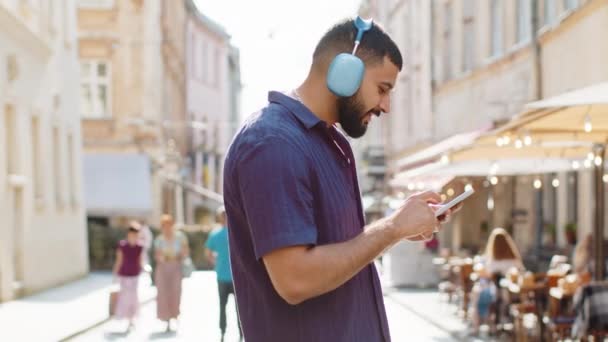 Χαρούμενη χαλαρή overjoyed indian άνθρωπος σε ασύρματα ακουστικά επιλέγοντας, ακούγοντας το αγαπημένο ενεργητική μουσική ντίσκο σε εξωτερικούς χώρους χορό smartphone. Guy τουριστικό περπάτημα περνά από αστική ηλιοφάνεια της πόλης δρόμο - Πλάνα, βίντεο