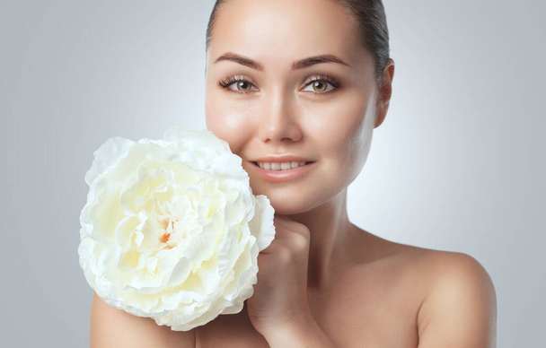 Porträt einer schönen, glücklichen Frau mit sauberer Haut. Sie hält eine wunderschöne weiße Pfingstrose in der Nähe ihres Gesichts.Kosmetologie Hautpflege und Make-up. - Foto, Bild