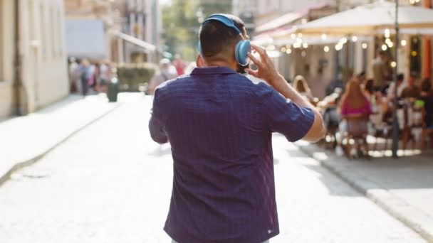 Vue arrière d'un touriste indien dans un casque sans fil marchant dans la rue à l'extérieur, écoutant de la musique, dansant, se tournant vers la caméra et souriant. Vue arrière de gars voyageur dans ville ensoleillée urbaine - Séquence, vidéo