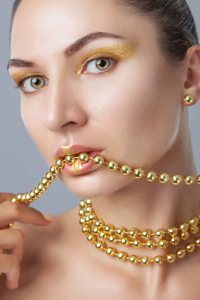 Portrait of a beautiful happy woman with beautiful creative makeup in gold colors. She has gold beads on her neck, in her hands and on her lips, and gold earrings. Make-up concept. - Photo, Image