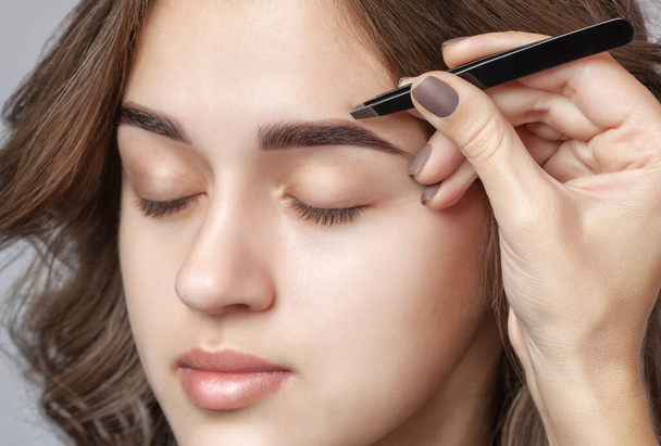Make-up artist plucks eyebrows with tweezers to a woman with curly brown hair and nude make-up. Beautiful thick eyebrows close up. Professional makeup and cosmetology skin care. - Photo, Image