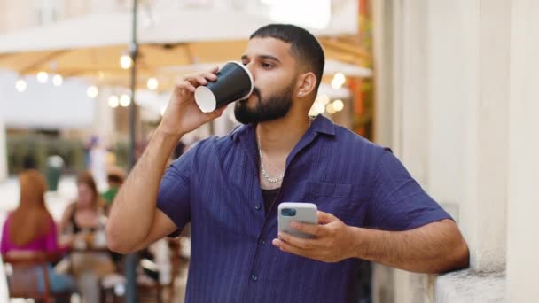 Heureux homme indien barbu dégustant le café du matin boisson chaude et souriant à l'extérieur. Détendant, faisant une pause. Un type marchant dans la rue urbaine ensoleillée, buvant du café. Mode de vie en ville à l'extérieur - Séquence, vidéo