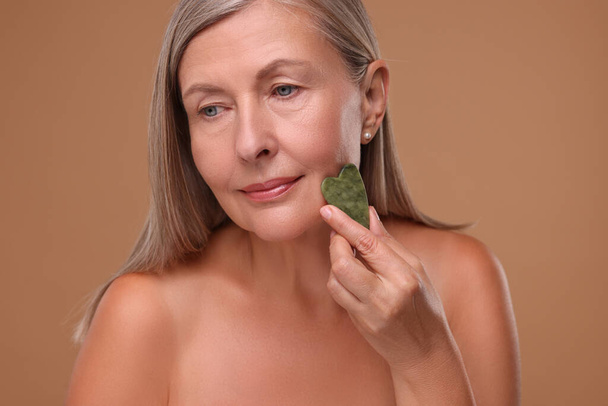 Woman massaging her face with jade gua sha tool on brown background - Foto, imagen