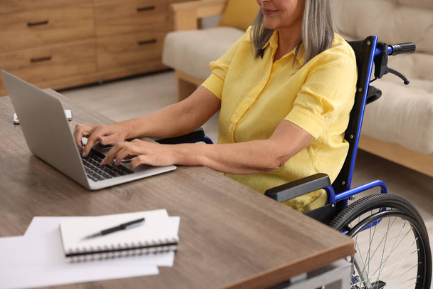 Woman in wheelchair using laptop at table indoors, closeup - Photo, image