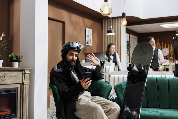 Καυκάσιος καλεσμένος κάθεται στον καναπέ περικυκλωμένος από εξοπλισμό σνόουμπορντ, έχοντας βιντεοκλήση στο ψηφιακό του τηλέφωνο. Tourist in snow clothing on videoconference with mobile device in ski resort σαλόνι. - Φωτογραφία, εικόνα