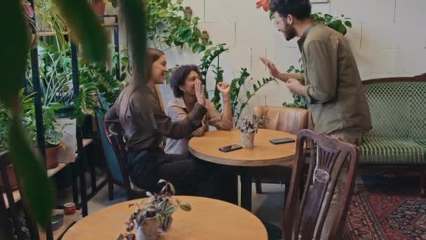 Yeşil saksı bitkilerinin arasında rahat bir kafede oturan iki farklı kızın orta boy fotoğrafı. Genç Afrikalı Amerikalı erkek arkadaş içeri giriyor, beşlik çakıyor ve onlara katılıyor. - Video, Çekim