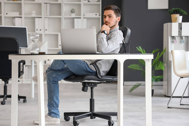 Красивый молодой человек сидит и работает с ноутбуком в офисе - Фото, изображение