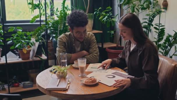 Orta boy beyaz bir kadın ve Orta Doğulu bir adamın rahat yeşil bir kafede oturup kahve içip hamur işi yerken pazar araştırması ve yeni iş girişimlerini tartışırken çekilmiş. - Video, Çekim