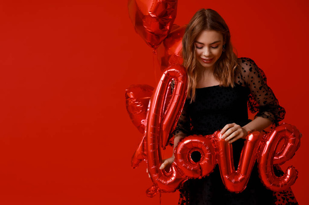 Νεαρή γυναίκα με λέξη ΑΓΑΠΗ φτιαγμένη από μπαλόνια σε κόκκινο φόντο. Γιορτή του Αγίου Βαλεντίνου - Φωτογραφία, εικόνα