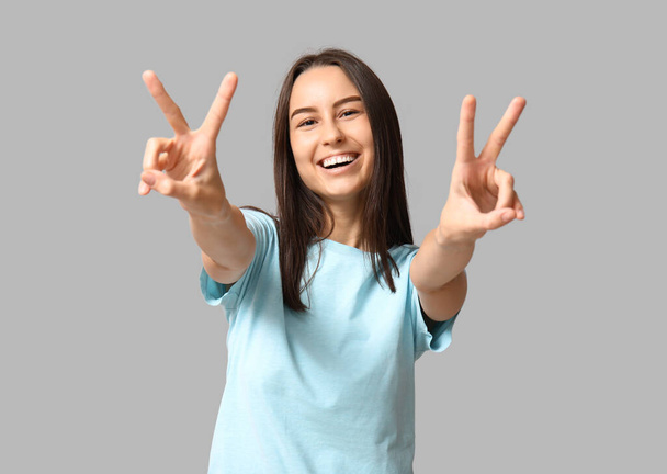 Красивая молодая женщина показывает победный жест на сером фоне - Фото, изображение