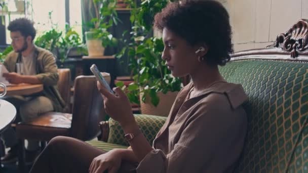 Foto lateral media de la joven mujer afroamericana ocupada con auriculares inalámbricos sentados en el sofá en la oficina o coworking, escribiendo mensaje o correo electrónico en el teléfono inteligente, un montón de plantas verdes en el fondo - Imágenes, Vídeo