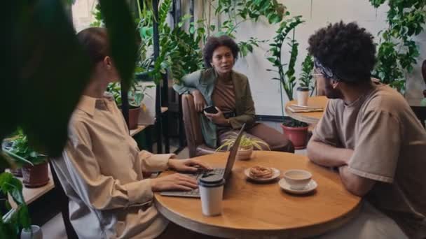 Média foto de mulher caucasiana com laptop e homem biracial sentado à mesa no escritório cantina ou co-working e conversando com colega afro-americano do sexo feminino na mesa ao lado - Filmagem, Vídeo