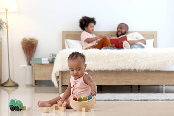 Afrikalı küçük şirin bebek bebek evde yatak odasında otururken oyuncak oynuyor. Babası ve kardeşi yatakta kitap okuyorlar. Mutlu aile yatak odasında birlikte vakit geçiriyor.. - Fotoğraf, Görsel