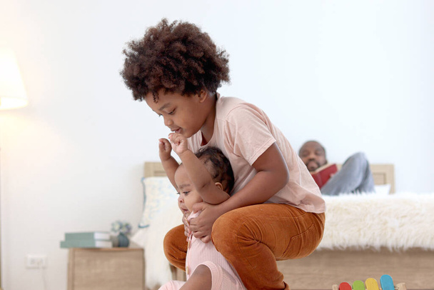 Heureux famille africaine passer du temps ensemble, frère garçon avec des cheveux bouclés noirs essayer de tenir et soulever petit bébé bébé bébé bébé sur le sol avec fond flou de papa lecture livre sur le lit. - Photo, image