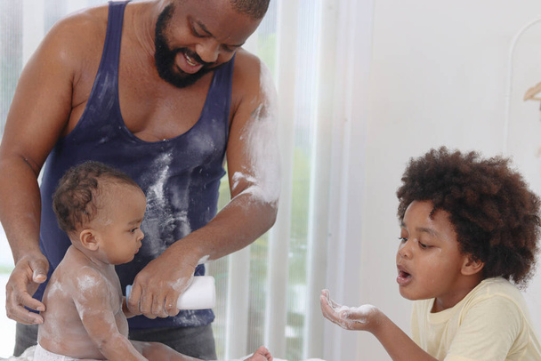 Happy African family, toddle baby infant sits on towel after takes a bath in bathroom, father and son brother hold dusting powder bottle and apply talcum powder on body of little kid daughter child. - Photo, Image
