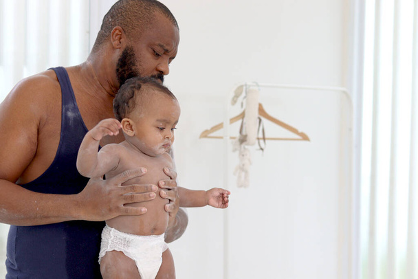 Happy African family, Father holding cute toddle baby infant kid after taking a bath and apply talcum powder on body ready in bathroom. Dad looks after little cute daughter child in family. - Photo, Image