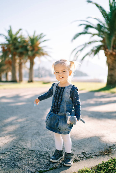 Malá usměvavá holčička stojí na cestě u palem s hračkou v ruce. Kvalitní fotografie - Fotografie, Obrázek