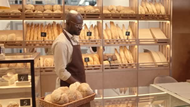Retrato de lentitud media de un joven calvo negro feliz trabajador de panadería en delantal parado en el mostrador y sonriendo a la cámara, vendiendo productos horneados frescos y postres - Metraje, vídeo