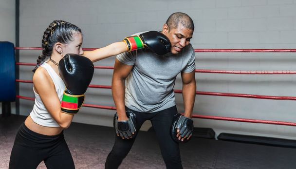 Horizontal photo pretty preteen caucasian girl with black gloves, black tights, white top and braids in her hair, practicing boxing with her black mid adult trainer, in the ring. Copy space. - Photo, Image