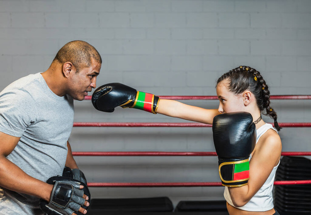 Horizontal photo pretty preteen caucasian girl with black gloves, black tights, white top and braids in her hair, with her black mid adult boxing trainer, in the ring. Sport, recreation concept. - Photo, Image