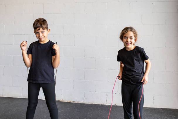 Горизонтальні фото елементарного хлопчика і дівчинки, одягнені в чорний спортивний одяг, посміхаються стрибати з мотузками в спортзалі. Концепція спорту, відпочинок. - Фото, зображення
