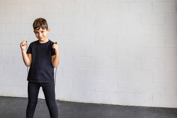 Horizontale foto elementaire jongen, gekleed in legging en zwart t-shirt, glimlachend springtouw in een sportschool. Begrepen, ruimte. Conceptsport, recreatie. - Foto, afbeelding