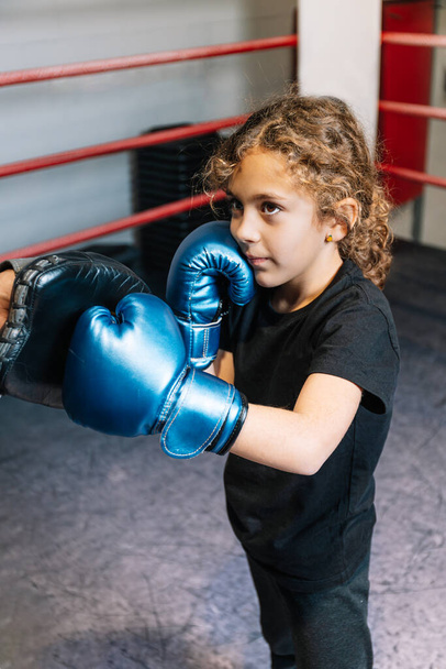 Függőleges fotó elemi lány, fekete pólóban, harisnyában és kék bokszkesztyűben, megtanul bokszolni a ringben a fekete középkorú boksz tanárával. Sport, szabadidő koncepció. - Fotó, kép