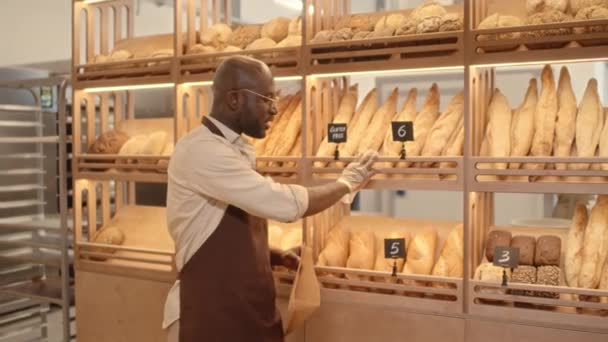 Medium shot van jonge Afro-Amerikaanse man in schort en wegwerp handschoenen zetten verse stokbrood in ambachtelijke papieren lange zak en verkopen aan blanke vrouwelijke klant staan bij de bakker teller - Video