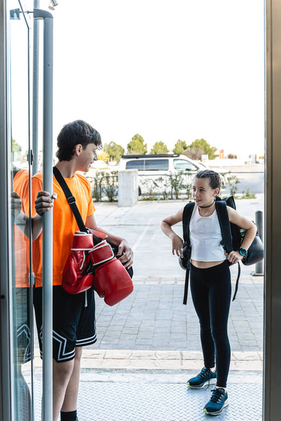 Vertikální foto chlapec dospívající a dospívající dívka přijíždějící do boxerské tělocvičny v dopoledních hodinách trénovat. Poslouchá, jak drží dveře, zatímco jí ustupuje. Sport, rekreační koncept. - Fotografie, Obrázek