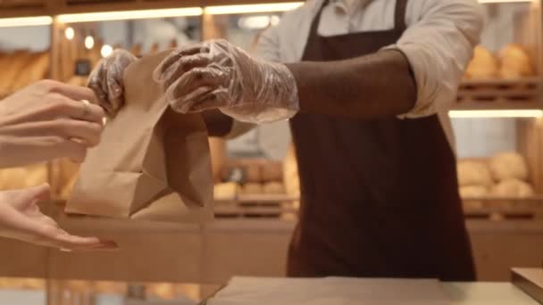 Καλλιεργημένη βραδύτητα του αγνώριστου μαύρου πωλητή φορώντας ποδιά και γάντια μιας χρήσης δίνοντας τσάντα χαρτί βιοτεχνίας στον πελάτη, ενώ στέκεται στον πάγκο στο άνετο αρτοποιείο - Πλάνα, βίντεο