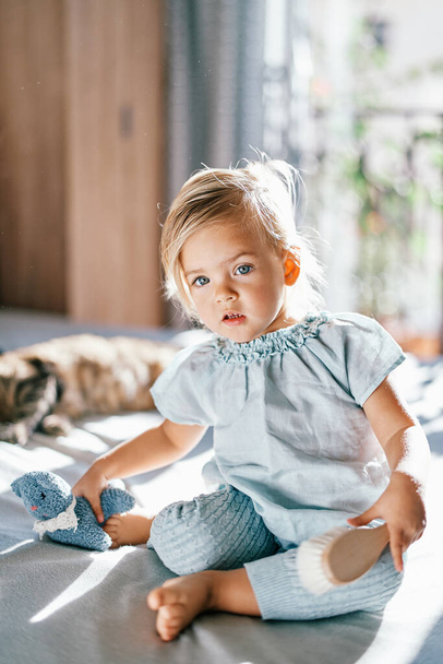 Little girl with a brush in her hand sits next to a soft toy on the bed. High quality photo - Photo, Image