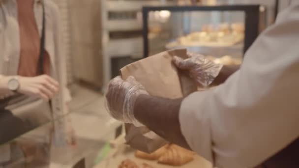 Przycinane powoli nierozpoznawalny sprzedawca rękawiczek ręcznych torba papierowa z torbą świeżego ciasta do klienta kobiety przy ladzie w małej przytulnej piekarni - Materiał filmowy, wideo