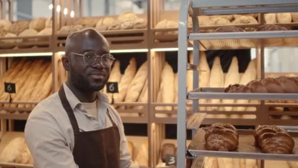 Cintura hacia arriba retrato del joven trabajador de panadería negro sonriente posando para la cámara con panes frescos en bandeja de aluminio - Metraje, vídeo